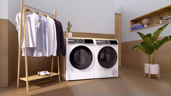 Eine Bosch-Waschmaschine und der passende Trockner stehen nebeneinander, links davon ein Holz-Kleiderständer mit mehreren T-Shirts, Hemden und Blusen.