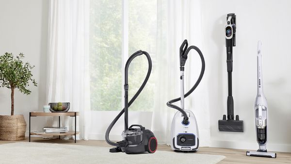 Vier Bosch-Staubsauger in Schwarz und Weiß (zwei kabelgebunden, zwei kabellos) stehen vor einer hellen Fensterfront mit weißen Vorhängen.