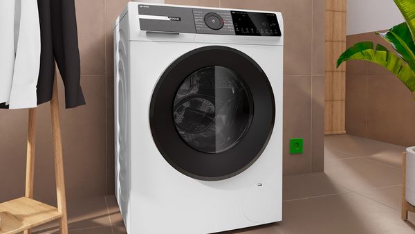 Bosch-Waschmaschine vor brauner Fliesenwand, daneben ein Kleiderständer mit zwei Hemden und eine Topfpflanze.