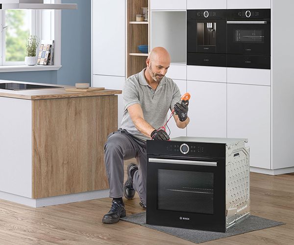 Egy f&eacute;rfi t&eacute;rdel egy fekete Bosch s&uuml;tő mellett, &eacute;s multim&eacute;terrel ellenőrzi a csatlakoz&aacute;sokat egy modern konyhai k&ouml;rnyezetben.