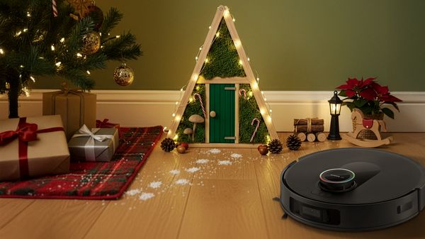 Auf einem Laminatfußboden steht neben einem Weihnachtsbaum ein dekoriertes Wichtelhaus und daneben ein Bosch Saugroboter.