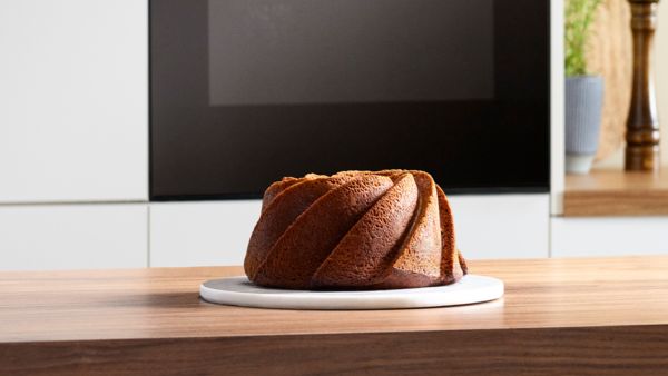Qızılı‑qəhvəyi bundt tort ağ boşqabda taxta masada, zərif m&uuml;asir mətbəx fonunda.