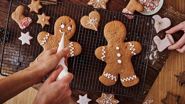 Eine Person verziert zwei Lebkuchenmännchen mit weißem Zuckerguss, die zusammen mit kleinen Lebkuchenherzen auf einem schwarzen Backgitter liegen.