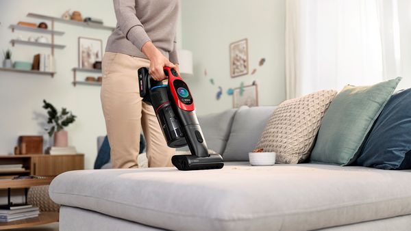 Eine Frau saugt mit dem Handteil eines Akku-Staubsaugers ein hellgraues Sofa ab.