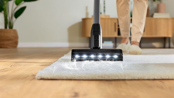 Blick von vorne auf die Düse eines Akku-Staubsaugers, mit dem eine Frau Tierhaare von einem Teppich saugt.
