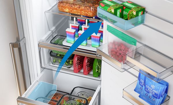 Прилежно организиран фризер, пълен със замразени ястия, плодове, зеленчуци и цветни ледени близалки, показва подвижна поставка на вратата, илюстрирана със синя стрелка от долния ляв към горния десен ъгъл за гъвкаво съхранение.