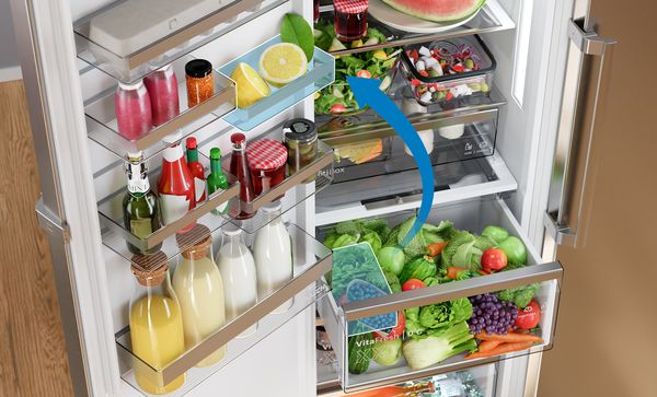 Добре организиран интериор на хладилник показва регулируеми рафтове и отделения на вратата, пълни с напитки, подправки, плодове и зеленчуци, със синя стрелка, подчертаваща гъвкавото разпределение за съхранение.