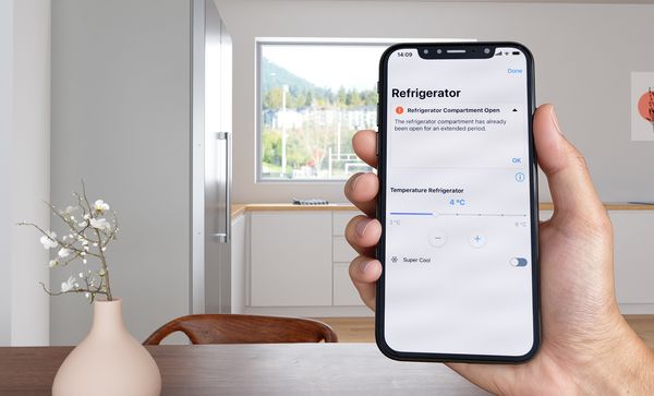 Ръка, държаща смартфон, показващ екрана на приложението Home Connect, докато на заден план се вижда модерна кухня с хладилник от неръждаема стомана.