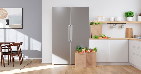 Врати на хладилник и фризер Single Door една до друга в светла кухня, заобиколени от хартиени пазарски чанти, пълни с пресни продукти.