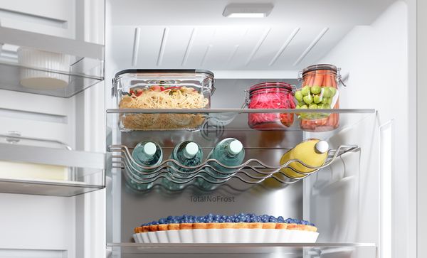 Изображението показва ярко осветен интериор на хладилник със стъклени буркани, бутилирани напитки във вълнообразна метална поставка и тарт с боровинки на долния рафт, всички осветени от вградена LED светлина.