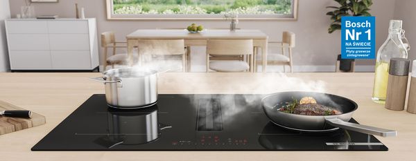 Płyta grzewcza zintegrowana z wyciągiem marki Bosch. Patelnia ze stekiem i garnek na płycie grzewczej, w tle jadalnia ze stołem.