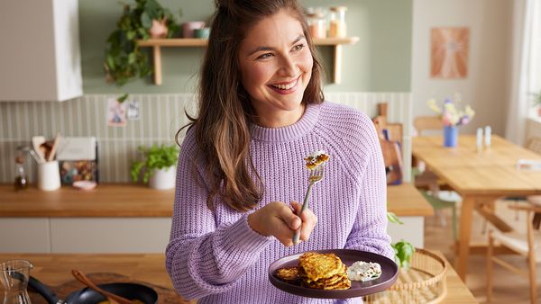 Eine Frau mit fliederfarbenem Pullover steht in einer hell eingerichteten K&uuml;che und isst ein Broke-Gericht mit der Gabel von einem Teller.