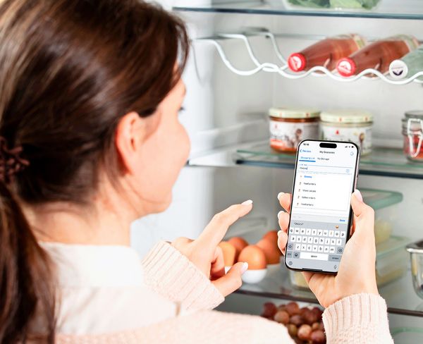 En person står framför ett öppet kylskåp, håller i en smartphone och går igenom en inköpslista.
