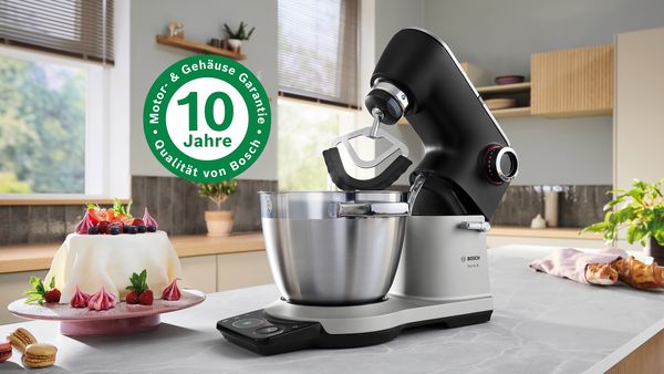 Eine Bosch K&uuml;chenmaschine Serie&nbsp;8 in Schwarz und Silber steht auf einer K&uuml;chenarbeitsplatte. Eine Plakette zeigt eine &bdquo;Motor- und Geh&auml;usegarantie f&uuml;r 10&nbsp;Jahre&ldquo;. Ein dekorierter Kuchen und Macarons stehen daneben.