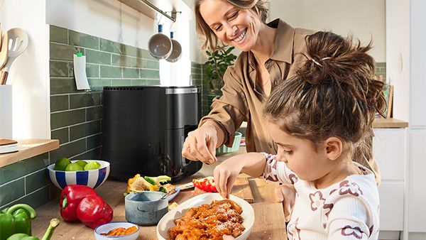 In einer hellen, modern gestalteten K&uuml;che stehen Mutter und Tochter an einer K&uuml;chenzeile und bereiten gemeinsam mit frischen Zutaten leckere Gerichte zu, die mit dem schwarzen Bosch Air Fryer zubereitet werden.
