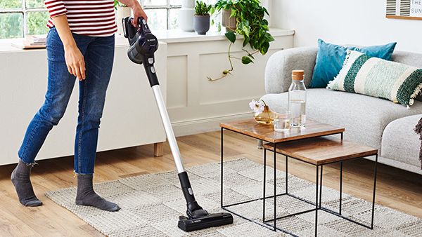 Es ist der Unterkörper einer Person zu sehen, die Jeans und ein rot-weiß gestreiftes Langarmshirt trägt und mit einem kabellosen Staubsauger von Bosch einen Teppich in einem hellen Wohnzimmer saugt.
