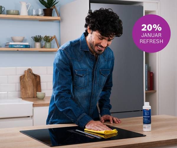 Una persona in camicia di jeans pulisce un piano cottura in vetroceramica nero su un'isola da cucina in legno con un panno giallo; a destra c'&egrave; un contenitore con un prodotto per la pulizia. Sullo sfondo una cucina moderna con mensole, piante, libri e un tagliere; in alto a destra un cerchio viola con la scritta "20% JANUAR REFRESH". 