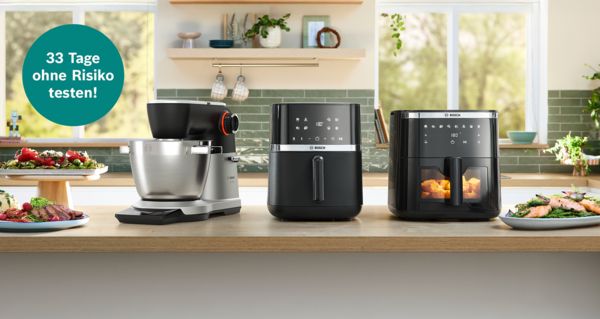 Bosch-K&uuml;chenmaschine und zwei Hei&szlig;luftfritteusen auf K&uuml;chentheke, daneben zubereitete Speisen.