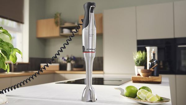 Una batidora de mano Bosch se encuentra sobre una encimera de cocina, con limas y hierbas en el fondo, mostrando un entorno de cocina moderno y elegante.