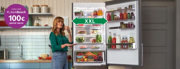 Žena stoji pored otvorenog hladnjaka ispunjenog zdravim namirnicama, prikazujući moderno kuhinjsko okruženje. 