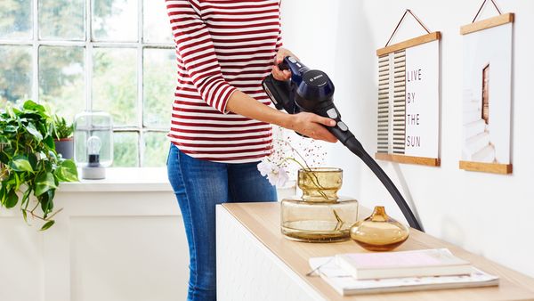 Es ist der Unterk&ouml;rper einer Person zu sehen, die Jeans und ein rot-wei&szlig; gestreiftes Langarmshirt tr&auml;gt und mit einem kabellosen Staubsauger von Bosch einen Teppich in einem hellen Wohnzimmer saugt.
