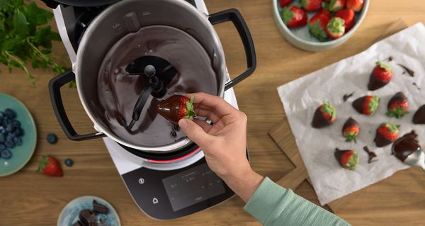 Cookit von Bosch beim Schmelzen von Schokolade &ndash; eine Hand taucht eine Erdbeere in fl&uuml;ssige Schokolade im Topf der K&uuml;chenmaschine.