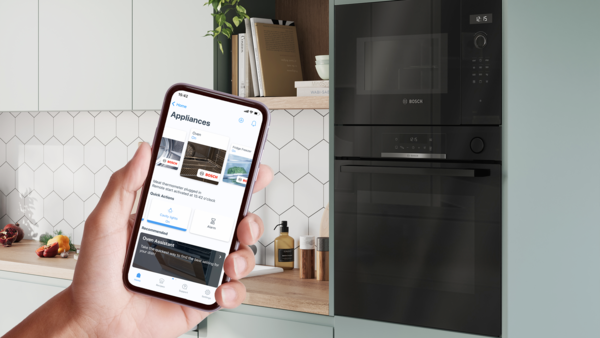 Əlində smartfon tutan şəxs; ekranda Bosch Home Connect tətbiqi a&ccedil;ıqdır və m&uuml;asir mətbəxdə soba ilə mikrodalğalı sobanı g&ouml;stərir.