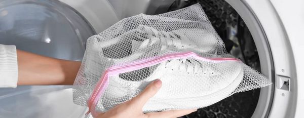 Mani che inseriscono nella lavatrice un paio di sneakers bianche all&rsquo;interno di un sacchetto in rete