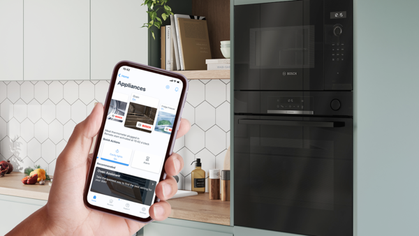 Əlində smartfon olan şəxs Home Connect tətbiqi vasitəsilə m&uuml;asir mətbəxdə quraşdırılmış Bosch sobasını və mikrodalğalı sobanı idarə edir.