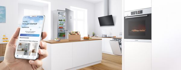 A hand holds a smartphone showing the contents of a fridge, which is visible in the bright kitchen in the background.