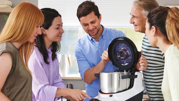 Zwei Frauen bereiten in einer hellen K&uuml;che mit einem modernen K&uuml;chenger&auml;t Lebensmittel zu, umgeben von frischen Zutaten und Kochutensilien