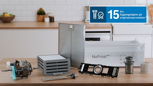 Et utvalg av originale reservedeler til hvitevarer, som en motor, et fettfilter, en NoFrost-skuff og et varmeelement, ligger p&aring; et trebord p&aring; et kj&oslash;kken. Et bl&aring;tt tekstbanner over bildet informerer om "15 &aring;rs tilgjengelighet p&aring; originalreservedeler".
