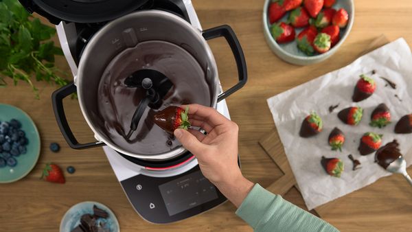 Cookit von Bosch beim Schmelzen von Schokolade &ndash; eine Hand taucht eine Erdbeere in fl&uuml;ssige Schokolade im Topf der K&uuml;chenmaschine.