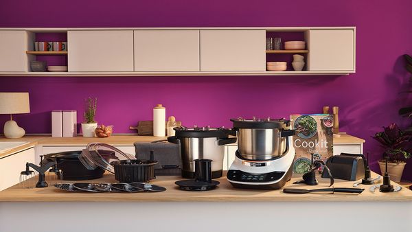Bosch Cookit K&uuml;chenmaschine mit Kochfunktion und komplettem Zubeh&ouml;r wie T&ouml;pfen, Werkzeugen, Scheiben und Kochbuch auf der Arbeitsfl&auml;che.