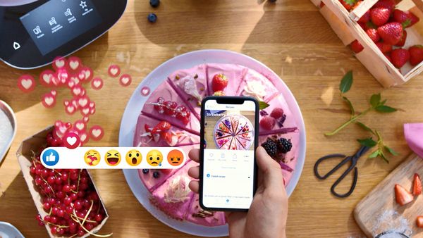 Smartphone mit Rezept in der Home Connect App &uuml;ber einer angerichteten Speise, umgeben von Zutaten und eingeblendeten Social-Media-Reaktionen.