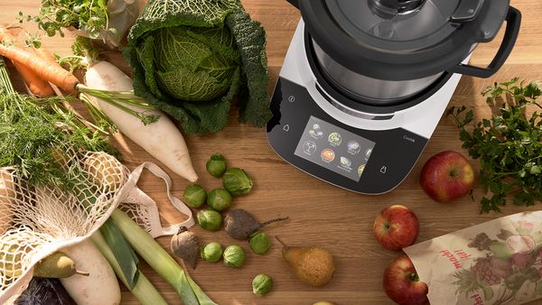 Cookit K&uuml;chenmaschine auf Holzarbeitsfl&auml;che, umgeben von frischem Gem&uuml;se, Kr&auml;utern, Pilzen, &Auml;pfeln und weiteren Zutaten zur Essensvorbereitung.