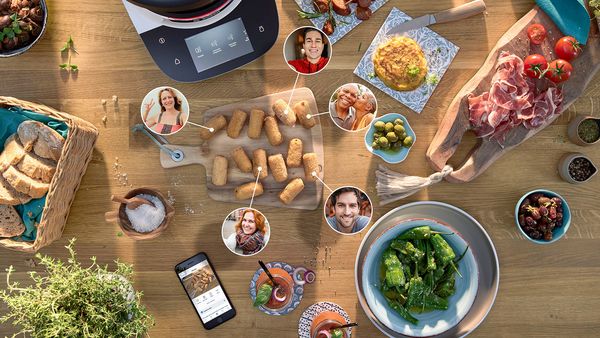 Cookit K&uuml;chenmaschine auf Holzarbeitsfl&auml;che mit verschiedenen Gerichten, Zutaten und eingeblendeten Profilbildern einer Community, die gemeinsam kocht und Rezepte teilt.