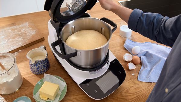 Hefeteig geht in der Bosch Cookit K&uuml;chenmaschine auf &ndash; Zutaten wie Mehl, Butter, Milch und Eier liegen auf einer Holzarbeitsfl&auml;che bereit.