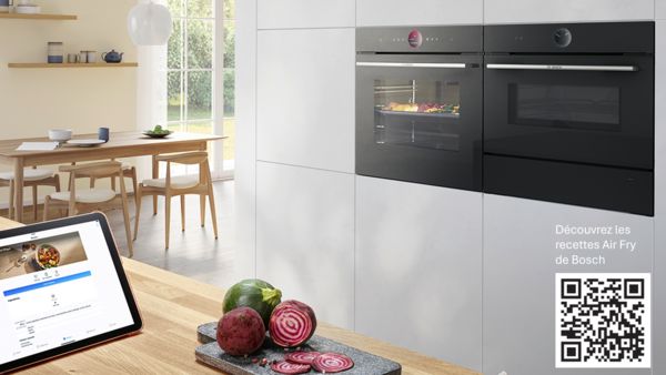 Cuisine &eacute;l&eacute;gante avec four int&eacute;grable S&eacute;rie 8 de Bosch avec fonction Air Fry et tiroir culinaire chauffant. Cuisson de diff&eacute;rents l&eacute;gumes au four. Plus de l&eacute;gumes sur une planche &agrave; d&eacute;couper d&eacute;pos&eacute;e sur un comptoir.