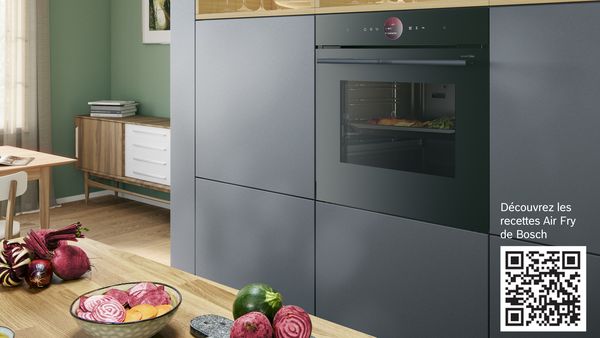 Cuisine &eacute;l&eacute;gante avec un four encastrable Bosch Series 8 de la gamme accent, &eacute;quip&eacute; de la fonction Air Fry et d&rsquo;un tiroir de maintien au chaud. Divers l&eacute;gumes cuisent dans le four, tandis que d&rsquo;autres l&eacute;gumes sont pos&eacute;s sur une planche &agrave; d&eacute;couper sur le plan de travail.