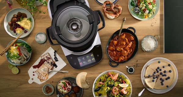 Bosch Cookit K&uuml;chenmaschine auf einer Holzarbeitsfl&auml;che, umgeben von vielf&auml;ltigen Gerichten wie Curry, Reis, Pasta, Kuchen, Waffeln und Salaten &ndash; Inspiration f&uuml;r die Cookit Rezeptewelt mit unterschiedlichen Kocharten.