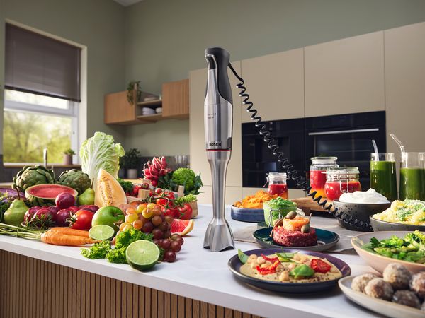Ein Stabmixer mit einem Spiralkabel steht auf einer Arbeitsplatte in der K&uuml;che umgeben von viel Obst, Gem&uuml;se, S&auml;ften, Suppen und Dips.
