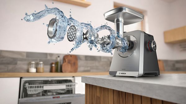 Месомелачка MultiPower Серия 8 на кухненски плот; нейните приставки и компоненти плуват в поток от вода пред уреда и отворена съдомиялна машина, видима на заден план.