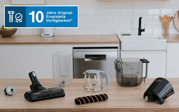 Auf einer K&uuml;chenarbeitsplatte liegen verschiedene Bosch‑K&uuml;chenger&auml;tekomponenten; ein Banner weist auf 10 Jahre Verf&uuml;gbarkeit originaler Ersatzteile hin.