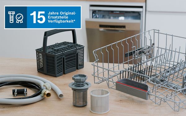 Geschirrsp&uuml;ler‑Ersatzteile auf einer Holzoberfl&auml;che, darunter Besteckkorb, graues Gestell, Schl&auml;uche und Filter, mit einem Banner zur 15‑j&auml;hrigen Verf&uuml;gbarkeit von Ersatzteilen.