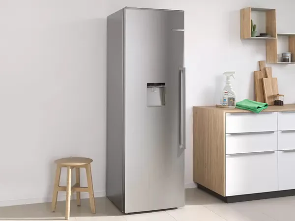 Tall stainless fridge next to a wooden cabinet; Bosch Fridge Cleaner spray bottle and a green cloth sit on the countertop.