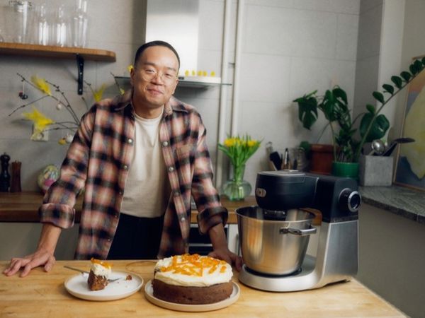 En person st&aring;r i et k&oslash;kken med en kage toppet med appelsinskiver og en skive serveret p&aring; en tallerken. En r&oslash;remaskine er i n&aelig;rheden med friske blomster i baggr