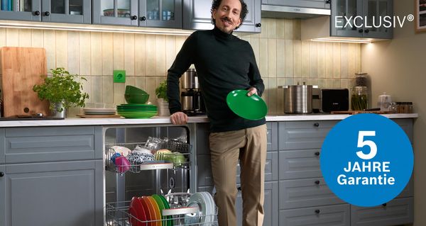 Person in moderner K&uuml;che l&auml;dt Geschirr in Geschirrsp&uuml;ler mit '5 Jahre Garantie'.