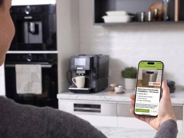 Person h&auml;lt Smartphone mit Produktanzeige eines Geschirrsp&uuml;lers in moderner K&uuml;che.