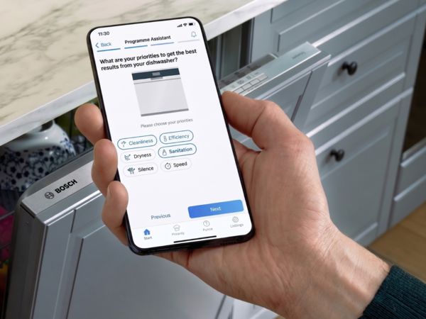 Hand h&auml;lt Smartphone mit Programm-Assistenten neben offenem Bosch-Geschirrsp&uuml;ler in moderner K&uuml;che.
